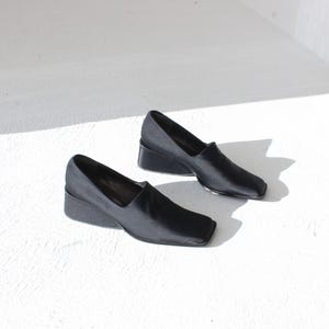 May include: A pair of black leather slip-on shoes with a square toe and a low, angled heel. The shoes are set against a white background, with natural light casting shadows.