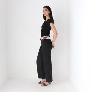 May include: A woman wearing a black short-sleeved shirt with a white belt and black pants. She is standing in a white room with her hands on her hips.