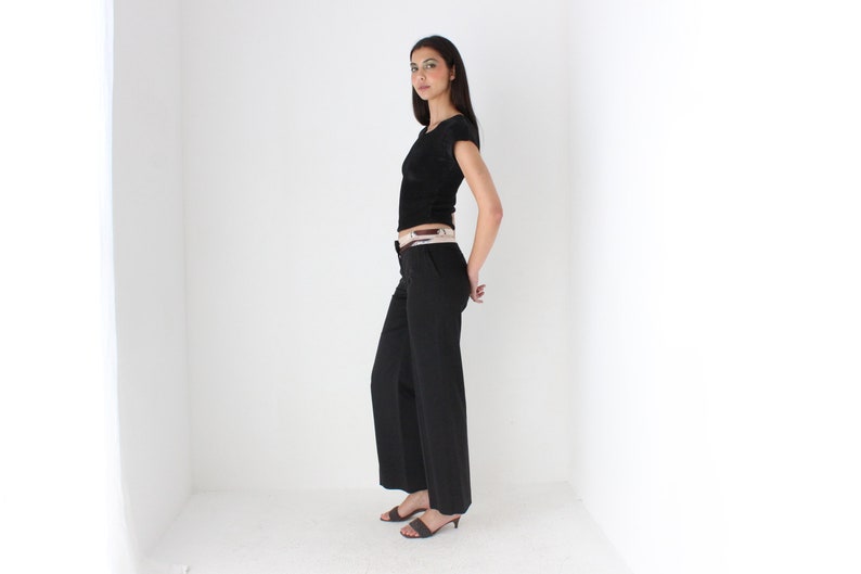 May include: A woman wearing a black short-sleeved shirt with a white belt and black pants. She is standing in a white room with her hands on her hips.
