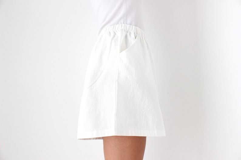 May include: White skirt with an elastic waistband and a pocket. The skirt is made of a textured fabric and is knee-length. The model is wearing a white t-shirt.
