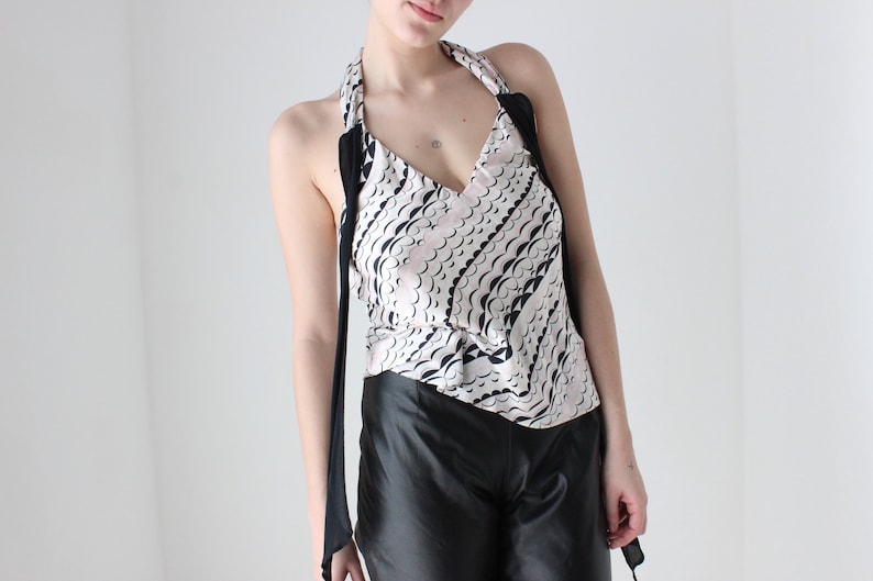 May include: A white halter top with a black and pink abstract pattern. The top has a deep V-neckline and ties at the neck. The model is wearing black pants.