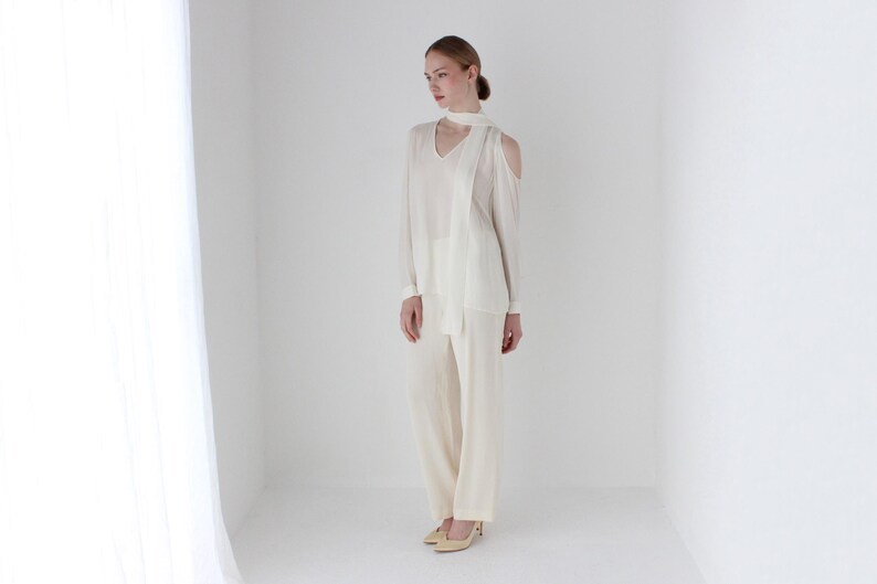 Puede incluir: Una blusa blanca de hombros descubiertos con una larga bufanda blanca atada alrededor del cuello. La blusa tiene mangas largas y un corte holgado. La modelo lleva pantalones blancos.
