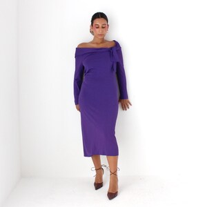 May include: A woman wearing a long-sleeved, purple off-the-shoulder dress with a bow detail at the neckline. She is wearing brown heeled shoes with laces.