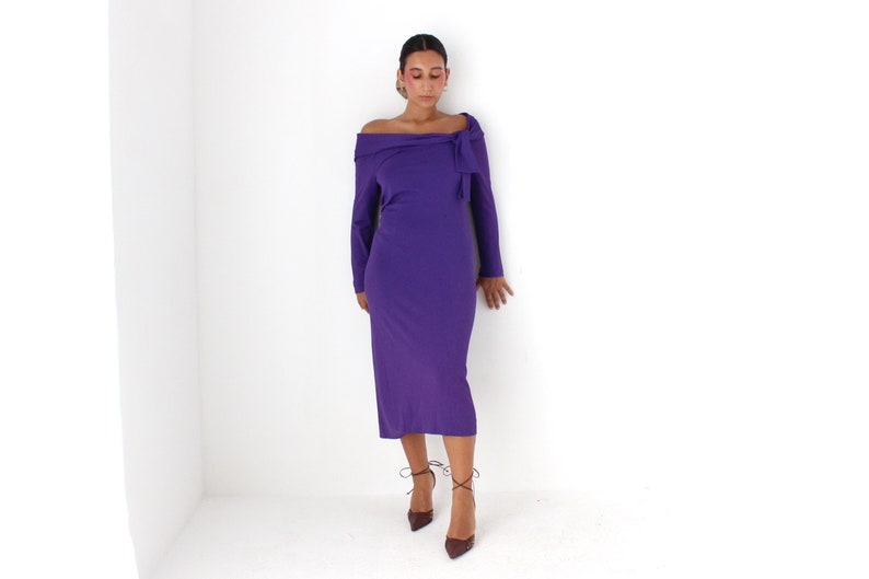 May include: A woman wearing a long-sleeved, purple off-the-shoulder dress with a bow detail at the neckline. She is wearing brown heeled shoes with laces.