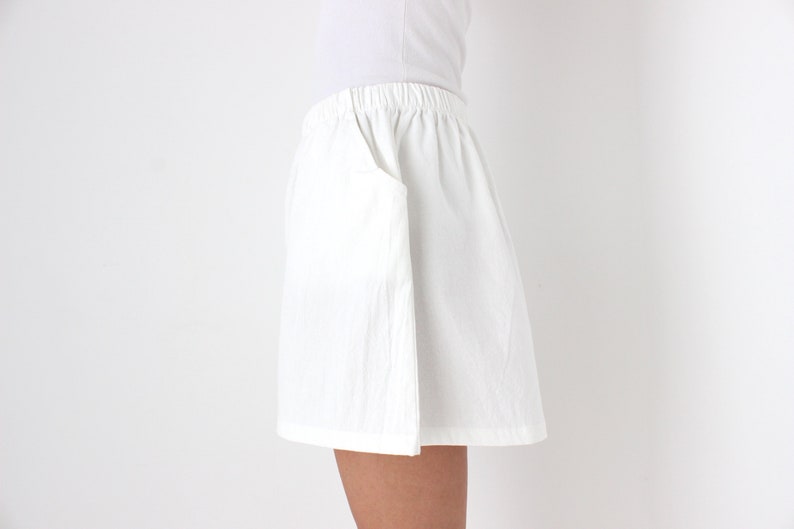 May include: White shorts with an elastic waistband and a side pocket. The shorts are made of a lightweight fabric and appear to be slightly textured. The garment is part of a casual clothing collection.