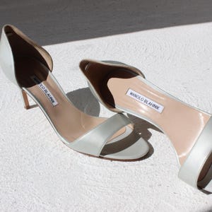 K&ouml;nnte beinhalten: Ein Paar hellgraue Satin-High-Heel-Sandalen. Die offenen Schuhe haben einen Riemen &uuml;ber den Zehen und einen d&uuml;nnen Absatz. Der Markenname "MANOLO BLAHNIK" ist auf der Innensohle sichtbar.