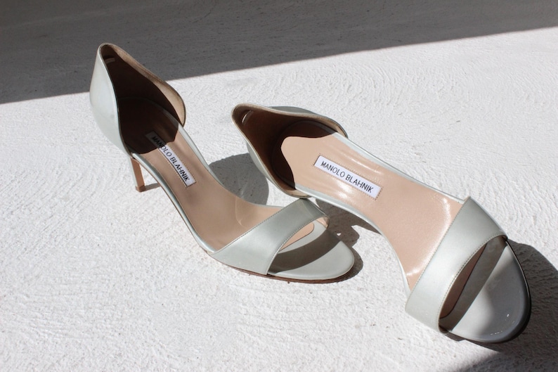 K&ouml;nnte beinhalten: Ein Paar hellgraue Satin-High-Heel-Sandalen. Die offenen Schuhe haben einen Riemen &uuml;ber den Zehen und einen d&uuml;nnen Absatz. Der Markenname "MANOLO BLAHNIK" ist auf der Innensohle sichtbar.