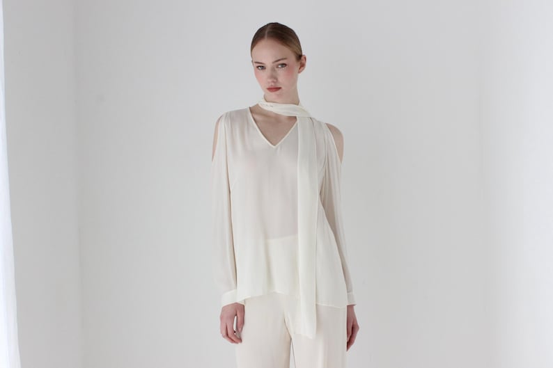 Puede incluir: Una blusa blanca de manga larga con cuello en V y una bufanda atada alrededor del cuello. La blusa tiene recortes en los hombros.