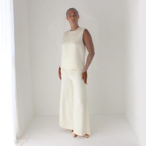 May include: A sleeveless ivory top and matching wide-leg pants. The outfit is made of a smooth, flowing fabric. A sheer veil is draped over the shoulders. The model is wearing gold-colored shoes. The ensemble is suitable for formal occasions.