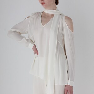 Puede incluir: Blusa blanca de manga larga con cuello en V y dise&ntilde;o de hombros descubiertos. La blusa tiene un lazo en el cuello.