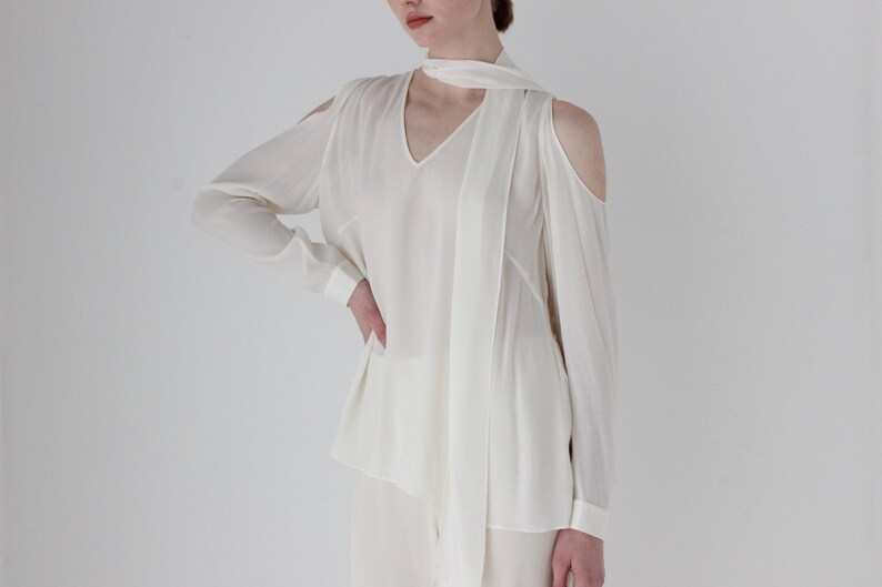 Puede incluir: Blusa blanca de manga larga con cuello en V y dise&ntilde;o de hombros descubiertos. La blusa tiene un lazo en el cuello.