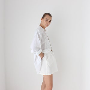 May include: A person wearing a white button-down shirt with rolled-up sleeves and white shorts. The outfit is simple and casual, with the shirt partially unbuttoned. The clothing is made of a light, breathable fabric.
