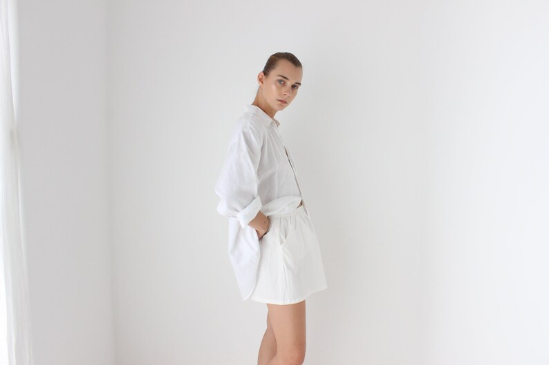 May include: A person wearing a white button-down shirt with rolled-up sleeves and white shorts. The outfit is simple and casual, with the shirt partially unbuttoned. The clothing is made of a light, breathable fabric.