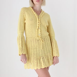 May include: A yellow crochet dress with long sleeves and a scalloped hem. The dress has a button closure at the neckline and a drawstring at the waist.