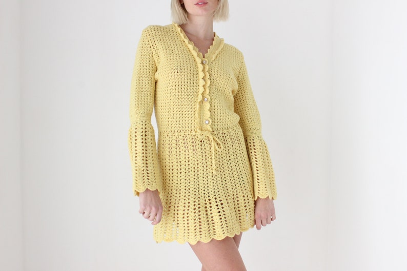 May include: A yellow crochet dress with long sleeves and a scalloped hem. The dress has a button closure at the neckline and a drawstring at the waist.