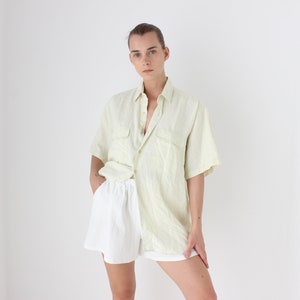 May include: A person wearing a light green short-sleeved button-down shirt and white shorts. The shirt has two chest pockets and is paired with white shorts. The outfit is casual and appears to be made of a lightweight fabric.