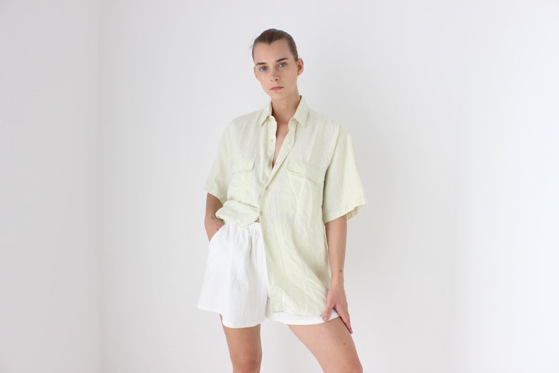 May include: A person wearing a light green short-sleeved button-down shirt and white shorts. The shirt has two chest pockets and is paired with white shorts. The outfit is casual and appears to be made of a lightweight fabric.