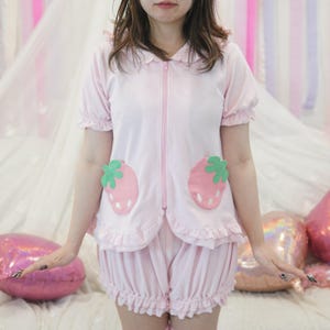 May include: A light pink pajama set featuring a zip-up top with two strawberry appliqués and matching ruffled shorts. The top has short sleeves and a Peter Pan collar. The pajamas are made of a soft, comfortable material.