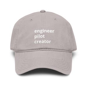 May include: A light gray baseball cap with the words "engineer pilot creator" printed in white. The cap has a curved brim and a six-panel design.