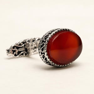 Può includere: Un gemello in argento con una pietra preziosa di corniola ovale, liscia e di colore rosso-arancio intenso, incastonata in una cornice metallica decorativa in stile filigrana con intricati motivi traforati e una finitura lucida.