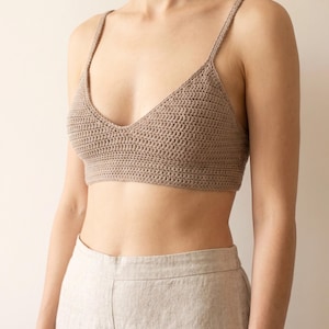May include: A light brown crochet bralette featuring a V-neckline, thin shoulder straps, and a horizontal ribbed band at the base. The garment is crafted with a dense, uniform stitch pattern and is paired with light beige linen-textured trousers.