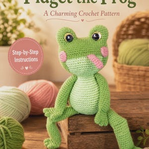 Może przedstawiać: Dokument ze wzorem szydełkowym zatytułowany Fidget the Frog A Charming Crochet Pattern z Step-by-Step Instructions przedstawiający zieloną szydełkowaną żabę z dużymi ciemnymi oczami, różowymi policzkami i różowym językiem, siedzącą na drewnianej skrzynce.