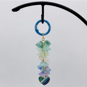 May include: A blue circular ring with white dots attached to a gold chain featuring a cluster of translucent bell-shaped flower charms in shades of green, white, and blue, ending with a small heart-shaped pendant with blue, green, and white stripes.