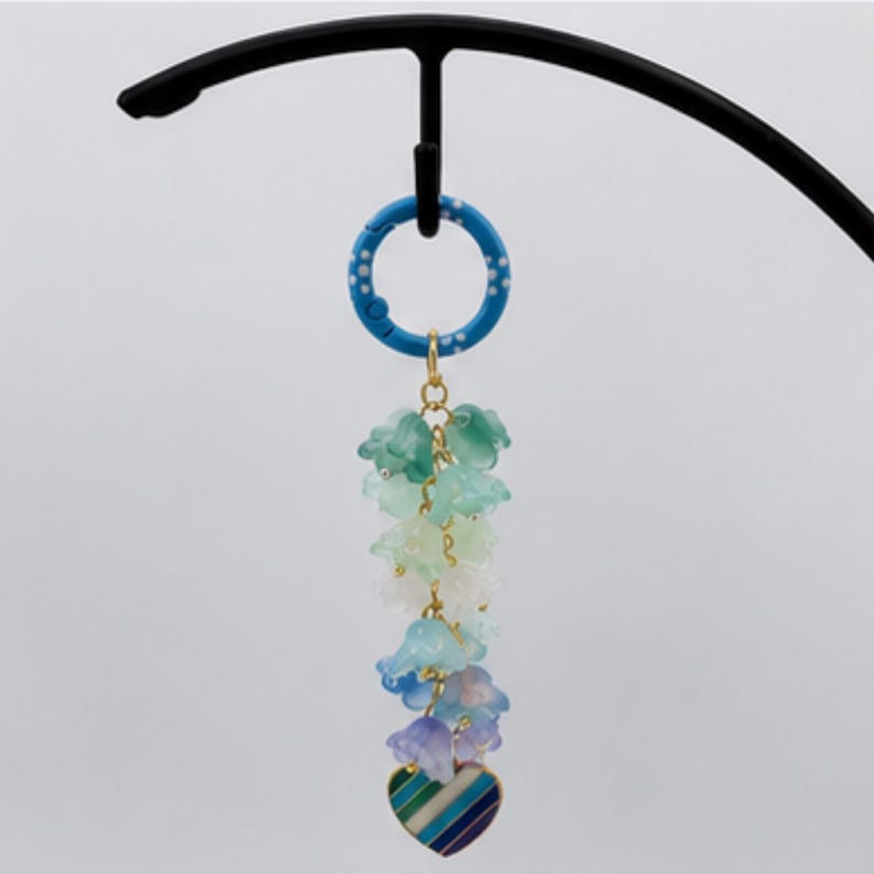 May include: A blue circular ring with white dots attached to a gold chain featuring a cluster of translucent bell-shaped flower charms in shades of green, white, and blue, ending with a small heart-shaped pendant with blue, green, and white stripes.