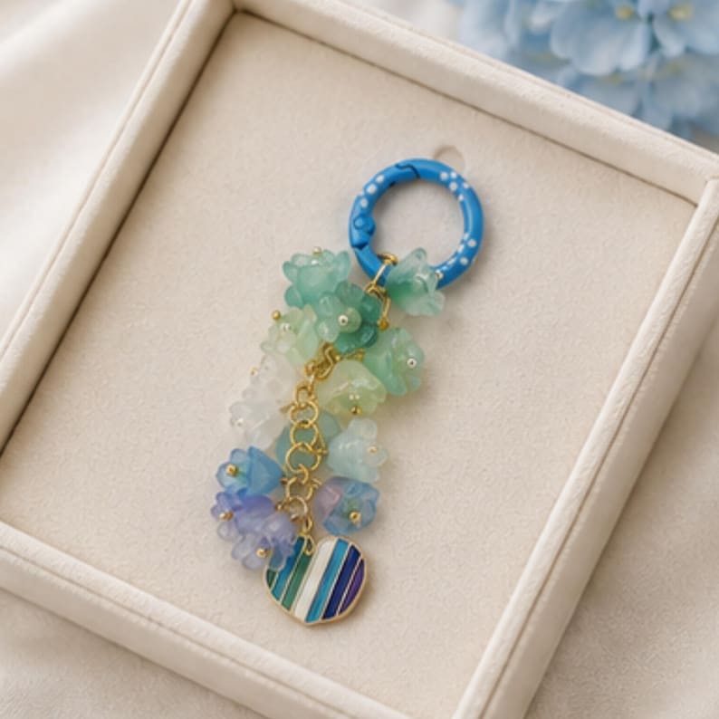 May include: A keychain featuring a blue circular ring with white dots, attached to a gold-toned chain adorned with multiple translucent flower-shaped beads in shades of green, blue, and purple, finished with a heart-shaped charm with blue stripes.
