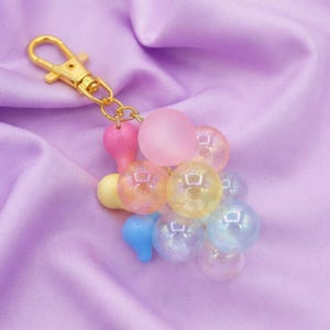 May include: A keychain featuring a gold-toned metal lobster clasp attached to a cluster of multi-colored, spherical beads. The beads include translucent, iridescent, and matte finishes in shades of pink, blue, yellow, and clear, creating a grape-like shape.