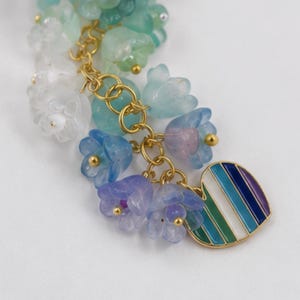 May include: A charm bracelet component featuring a cluster of translucent, bell-shaped flower beads in shades of white, mint green, light blue, and lavender. The beads are attached to gold-tone metal rings with a heart-shaped charm featuring vertical stripes of blue, green, and white enamel.