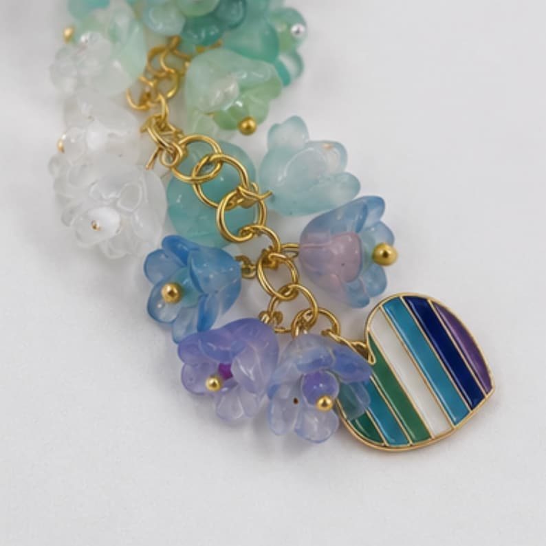 May include: A charm bracelet component featuring a cluster of translucent, bell-shaped flower beads in shades of white, mint green, light blue, and lavender. The beads are attached to gold-tone metal rings with a heart-shaped charm featuring vertical stripes of blue, green, and white enamel.