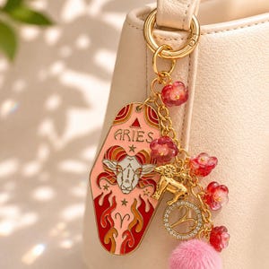 May include: Aries keychain with a pink and red design, featuring a ram illustration and the word "ARIES". The keychain includes a gold charm, pink pom-pom, and red flower accents, attached to a gold ring.