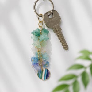 May include: A keychain featuring a gold-toned metal ring attached to a silver-toned key and a decorative charm. The charm consists of a gold chain adorned with small, translucent flower-shaped beads in a gradient of green, blue, and purple, ending in a heart-shaped pendant with vertical stripes in matching colors.