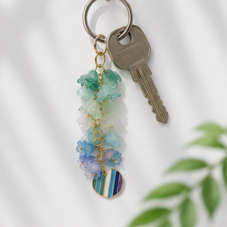 May include: A keychain featuring a gold-toned metal ring attached to a silver-toned key and a decorative charm. The charm consists of a gold chain adorned with small, translucent flower-shaped beads in a gradient of green, blue, and purple, ending in a heart-shaped pendant with vertical stripes in matching colors.