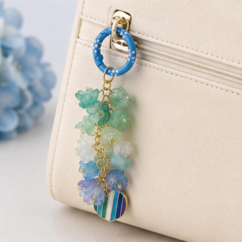 May include: A bag charm featuring a blue circular ring with white dots, attached to a gold-toned chain adorned with multiple translucent bell-shaped flower beads in shades of green, white, blue, and purple, ending with a heart-shaped striped charm.