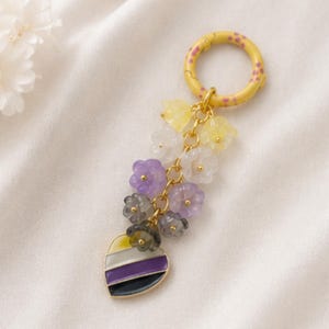 May include: A keychain featuring a yellow ring with pink spots, connected to a gold-tone chain with multiple small, translucent flower-shaped beads in yellow, white, purple, and gray, ending in a heart-shaped charm with yellow, white, purple, and black stripes.