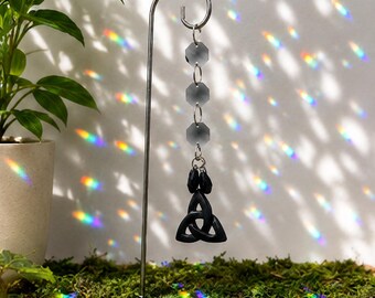 Pode incluir: Um suporte de gancho de metal prateado exibe um ornamento suspenso com três contas de vidro transparentes, facetadas e octogonais conectadas por anéis prateados, terminando em um pingente de triquetra preto com duas pequenas contas pretas.