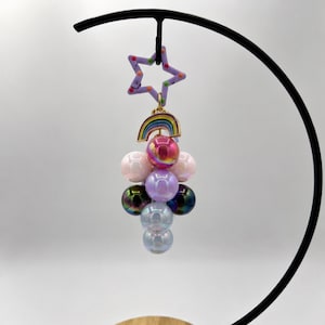 May include: A whimsical charm featuring a purple star with colorful polka dots and a smiling face, connected to a rainbow pendant and a cluster of iridescent and pastel-colored beads. The charm is suspended from a black metal stand.