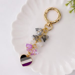 May include: A gold-toned metal keychain featuring a circular clasp and a dangling charm cluster. The cluster includes several small, translucent flower-shaped beads in shades of gray, white, and purple, ending in a heart-shaped charm with horizontal stripes.