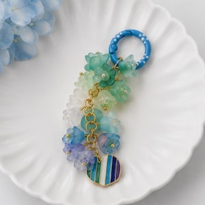 May include: A keychain featuring a blue circular ring with white dots, attached to a gold-toned chain adorned with multiple translucent flower-shaped beads in shades of green, blue, and white, ending with a heart-shaped charm with vertical stripes.