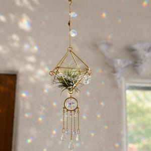 May include: A gold-toned geometric diamond-shaped metal frame hanging ornament containing a green air plant. The structure features clear faceted crystal beads and dangling chains with teardrop-shaped crystals that reflect light into rainbow patterns.