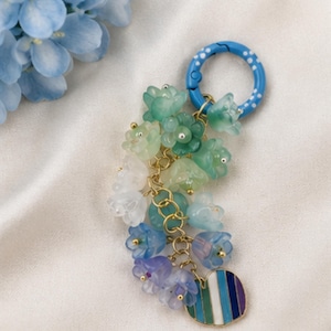 May include: A keychain featuring a blue ring with white polka dots, attached to a cluster of translucent flower-shaped charms in shades of green, white, blue, and purple, connected by gold-toned metal links, ending with a striped circular charm.
