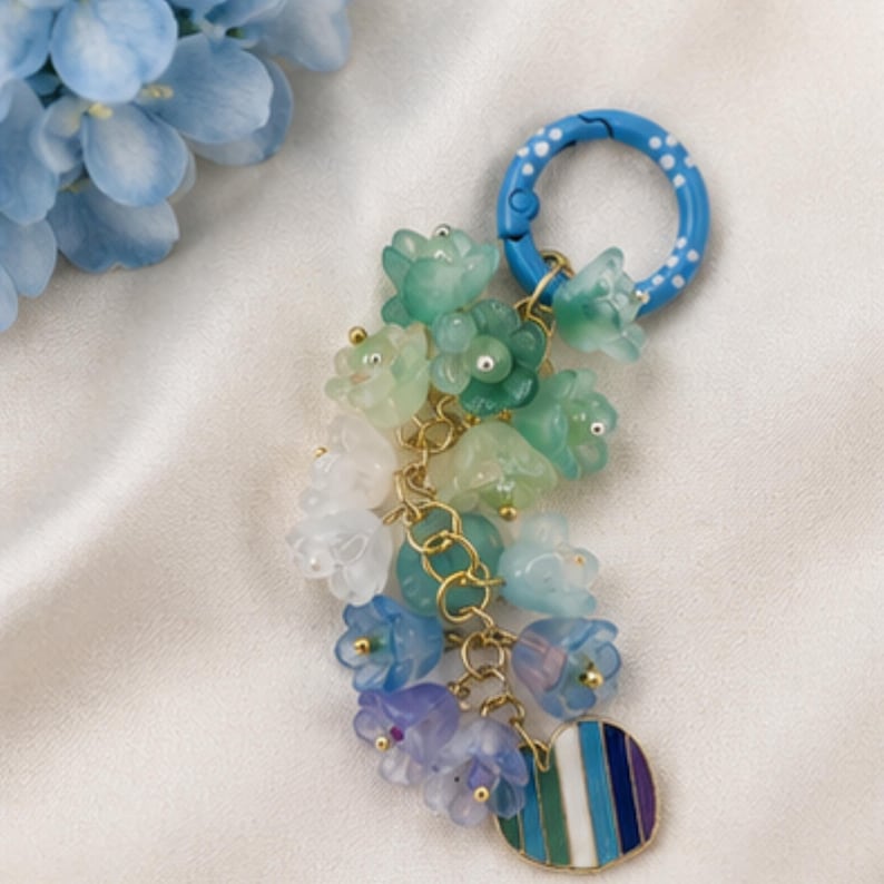 May include: A keychain featuring a blue ring with white polka dots, attached to a cluster of translucent flower-shaped charms in shades of green, white, blue, and purple, connected by gold-toned metal links, ending with a striped circular charm.