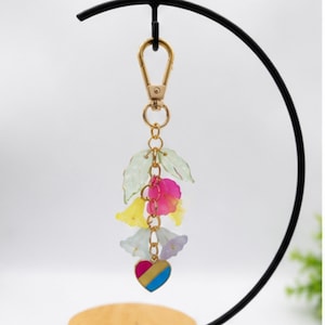 May include: A gold-toned metal keychain featuring a lobster clasp, a central chain, and several translucent, bell-shaped flower charms in shades of pale green, bright pink, yellow, and white, finished with a small heart-shaped charm with pink, yellow, and blue stripes.