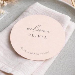 Può includere: Un segnaposto rotondo rosa chiaro con la parola "welcome" in corsivo, il nome "OLIVIA" in un font serif e la frase "We are so glad you're here!". Il segnaposto è su un tovagliolo di lino bianco.