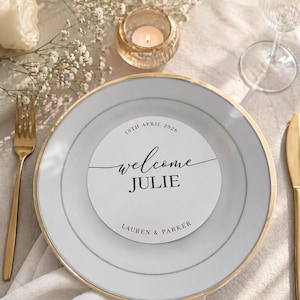 Puede incluir: Un plato blanco con borde dorado y una tarjeta circular que dice "welcome JULIE" con la fecha "10th April 2026" y los nombres "LAUREN & PARKER". Un tenedor y un cuchillo dorados están a cada lado del plato. Una vela encendida está en un portavelas de cristal.