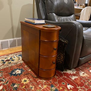 May include: A wooden side table with three drawers and a curved top. The table is a warm brown color and has a glossy finish. A book and a coaster sit on top of the table. The table is next to a leather recliner.