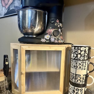 May include: A black stand mixer with floral accents and a stainless steel bowl sits above a small wooden cabinet with glass doors. Stacked black and white floral mugs are next to the cabinet. A black hand blender is on the left.