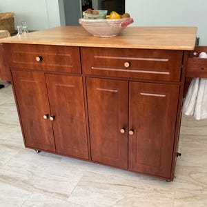 May include: A wooden kitchen island with a light-colored butcher block top, two drawers, and four cabinet doors. The unit features a rich brown finish, round knobs, and a side-mounted towel rack holding a white cloth. It sits on small wheels.